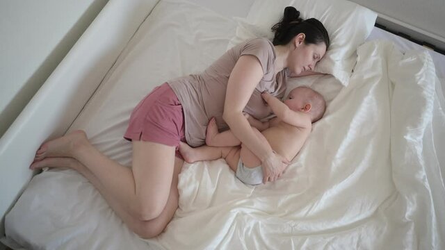Tired mother lying with newborn cute infant hyperactive baby boy on bed, holding him on arms, hugging and breastfeeds with breast milk. Healthy child, concept of hospital and happy motherhood. Nursery