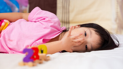 Child sleeps on the bed. Girl scratches his nose from the itch. Blur the wooden toys in front of the little kid. On a soft white mattress bed. Baby wearing a pink shirt is 4 years old.