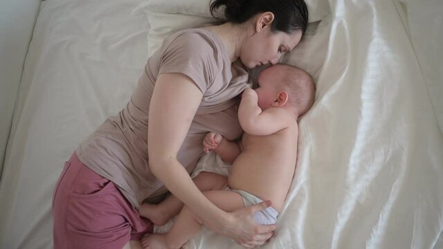 Young mother lying with newborn cute infant naked baby boy on bed, holding him on arms, hugging and breastfeeds with breast milk. Healthy child, concept of hospital and happy motherhood. Nursery.