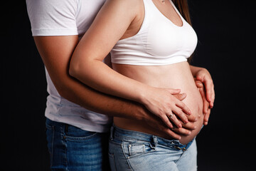 Husband and wife in white T-shirts and jeans on a black background hold hands on a pregnant belly. A pregnant woman with a man waiting for an unborn baby. On a black background without faces.