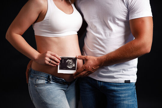 A man and a woman in white T-shirts and jeans on a black background are holding an ultrasound image of a baby on a bare pregnant belly. Loving couple awaiting the birth of a baby