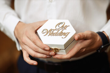 A man in a white shirt and blue trousers without a face with a smart watch holds in his hands a hexagonal wooden box with the inscription Our wedding. Groom with rings in a white case