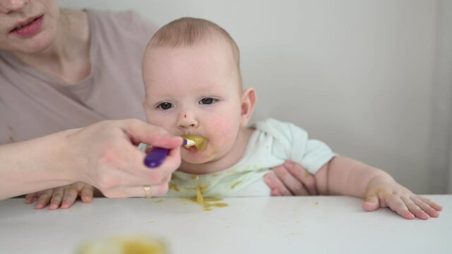 Little newborn funny baby boy learning to eat vegetable or fruit puree from glass jar with spoon. Young mother helping little son eat first food