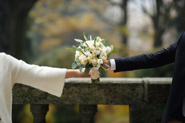 The bride and groom in a black tuxedo and a white fur coat on outstretched hands hold a wedding bouquet of peonies and white roses in nature among the forest. Man and woman without faces