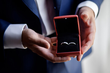 The man the groom in a white shirt and blue jacket holds on his palm wedding gold rings in a red velvet box by the window. Groom morning, close-up without a face only hands