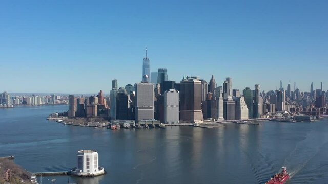 An aerial view of New York harbor on a sunny day with clear blue skies. The drone camera dolly in over the water, high enough to see Governors Island ventilation tower and Manhattan in the background.