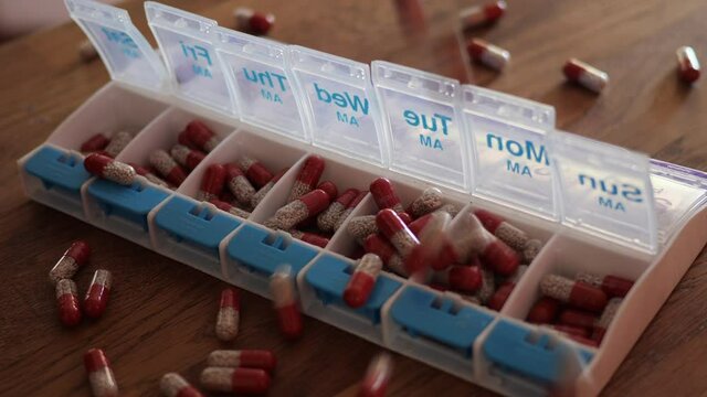 Pills falling on a weekly medication organizer container in slow motion, and onto a wood kitchen table. Creon for Cystic Fibrosis digestive enzyme treatment at home.