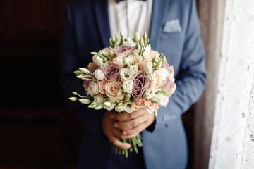 Man groom in a beautiful stylish classic suit holds a wedding bouquet of white and pink roses standing at the window waiting for the bride. Hands without a face.