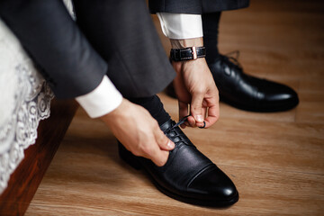 A strong man with his arms folded in a suit is tying shoelaces on black leather classic shoes. A groom with a ring on his hand in shoes for his wedding. Only hands without a face.