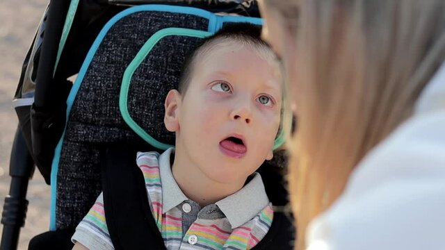 Close Up Little Kid Sitting In Special Wheelchair Outdoors. Child With Physical Disability Walking In The Park With Mom. Single Parent Caring Of Her Son