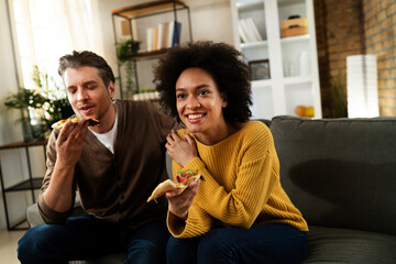 Cheerful young couple sitting on sofa at home. Happy woman and man eating pizza while watching a movie.