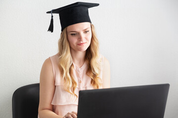 Woman student in masters hat takes an online exam, defends a diploma behind a laptop. Online education, graduation