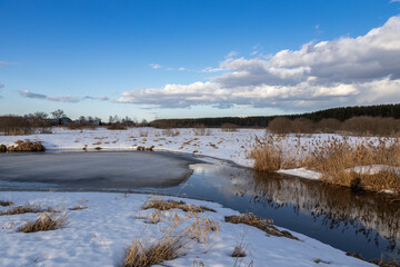 Early spring, blue sky and clouds are reflected in the river. Grass and snow in the foreground. Ice is melting on the river.