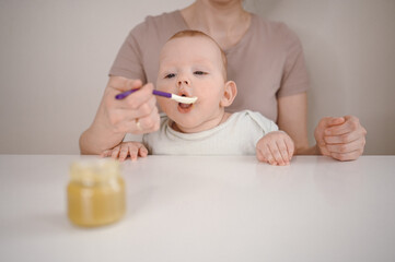 Obraz na płótnie Canvas Little newborn funny baby boy learning to eat vegetable or fruit puree from glass jar with spoon. Young mother helping little son eat first food