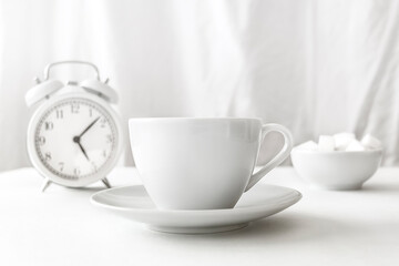 A cup of coffee, an alarm clock, and a sugar bowl are on the table. All items are white on a light background. White on white.