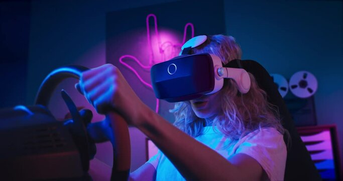 A girl is playing racing game in VR. She is sitting on the gaming chair with a steering wheel and pedals. A girl wears VR headset. Chair is located in the living room with red and blue neon lighting.