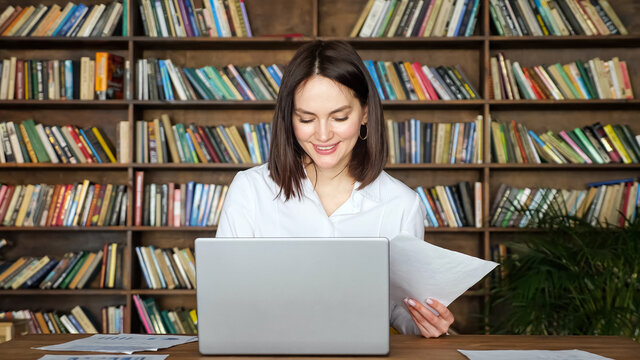 Smiling young woman in stylish white blouse works with papers near contemporary laptop at table against large library racks full of book