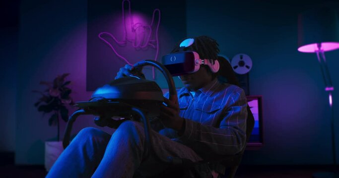 A guy is playing racing game in VR. He is sitting on the gaming chair with a steering wheel and pedals. A guy wears VR headset. Chair is located in the living room with red and blue neon lighting.