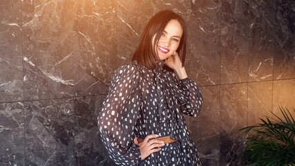 Joyful elegant brunette in long grey polka dot dress poses smiling and spins showing frock near marble wall, sunlight