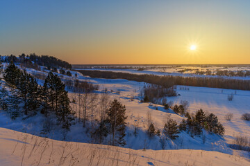 Sunset over the Irtysh river valley in Siberia (Russia) on a cold winter evening. Bird's eye view of the frozen river. There are many trees and vegetation along the banks. Blue-yellow sky. Rainbow sun