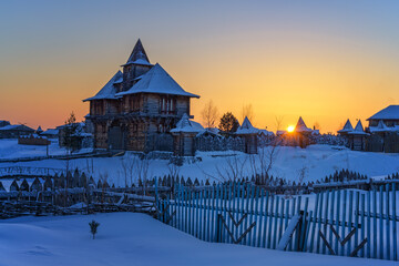 Sunset over a Siberian village (Russia) on a cold winter evening. Wooden log houses with a fence and triangular turrets against a blue sunset sky with yellow-orange paints. Lots of white snow  © olgaS