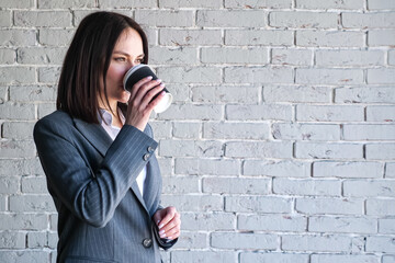 Fototapeta na wymiar Smiling company manager in white blouse and grey classic costume drinks coffee from paper cup standing against brick wall before work copyspace