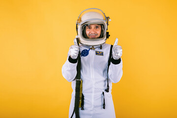 Male cosmonaut in spacesuit and helmet, raising thumbs, on yellow background.