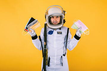 Male cosmonaut in spacesuit and helmet, holding many colored surgical masks, on yellow background. Covid-19 and virus concept