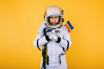 Gay transsexual male cosmonaut with serious gesture in space suit and helmet, holding lgtbi rainbow flag and transgender flag, on yellow background.