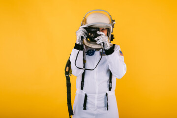 Male cosmonaut in space suit and helmet, taking a picture with a retro camera, on yellow background.