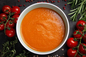 Top view of a bowl of tomato cream soup. The ingredients are arranged around the bowl: cherry tomato twigs, rosemary and thyme twigs, pink pepper and salt