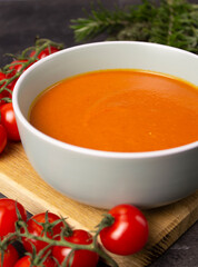 Rich color tomato puree soup in a gray bowl on a wooden board with cherry tomatoes and herbs on a background