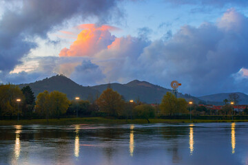 Scenic landscape view of a river with a windmill next to the mountains during the blue hour in the dawn, Limia River, Ponte de Lima, Portugal