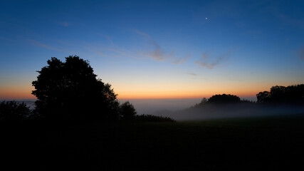 Sunrise at dawn, foliage trees in the fog. Blue sky with soft orange tones, the moon and the planet Venus can be seen. Copy space. Germany, Swabian Alb.