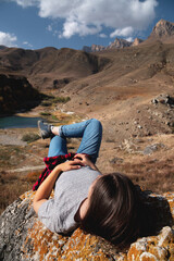 young happy woman lies on stones and looks at rocky mountains and lake, sunny day