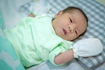 A close-up picture of an Asian baby lying on the mattress. She was wearing a green shirt and gloves.