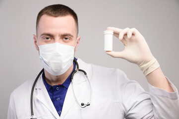 Young caucasian male medical doctor in a protective mask with a white can of pills in his hand shows it while looking into the camera. Advertising tablets of vitamins and dietary supplements