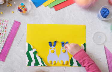 Child making card with Easter bunnies toys from colorful paper and placticine. Handmade. Project of children's creativity, handicrafts, crafts for kids.