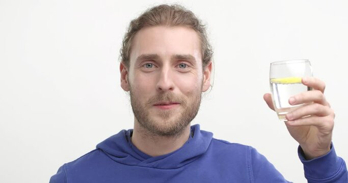 Happy bearded Young Man Drinking water with lemon from glass on White Background. Showing Healthy Lifestyle. Standing at studio wearing Blue Sweatshirt. Having Healthy Skin. Life. Water Balance.