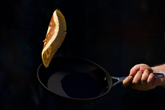 Hand holding a black frying pan and flipping a pancake in the air against a black background