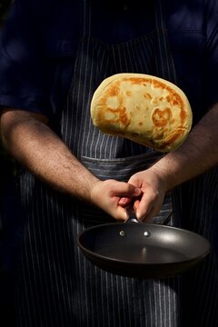 A chef in a pin striped apron using both of his hands to flip a pancake in the air against a dark black background