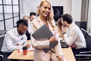 Confident young woman in formal clothes, smiling with laptop in hads and looking on the camera sitting on the edge of the table in the office