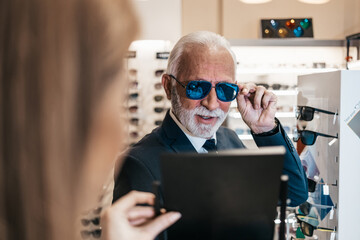 Elegant senior businessman choosing and buying sunglasses in optical store and young female seller...
