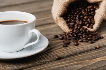 blurred sack bag with roasted coffee beans near cup on wooden surface