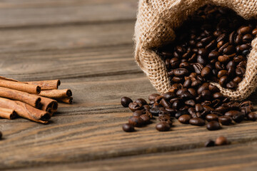 Sack bag with roasted coffee beans near cinnamon sticks on blurred wooden surface