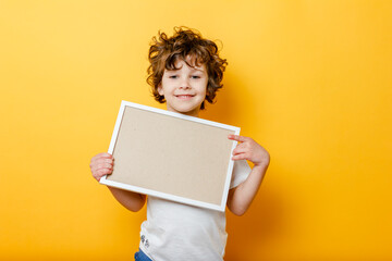 curly cute boy in white T-shirt holds an empty frame for text, smiling and shows up finger on bright yellow background.white signboard or banner - copy space concept to fill text. Message board