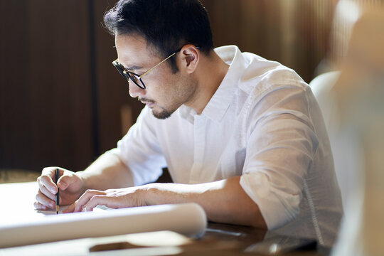 Asian bearded designer sitting, thinking and sketching ideas indoors