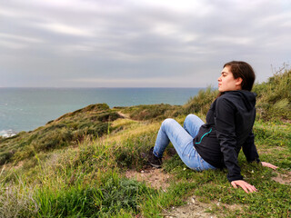 Naklejka na ściany i meble Young woman sitting on some rocks, on top of some dunes, watching the sea.
