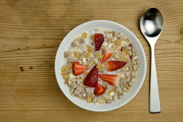 Healthy breakfast concept with oat flakes and fresh straw berries with milkon rustic wooden table with steel spoon beside a white bowl.