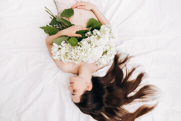 Charming attractive pregnant young female gently holds hands on tummy lying on comfy white bed at...